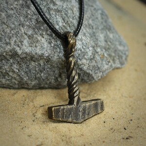May include: A bronze-colored Mjolnir pendant necklace with a twisted handle, hanging on a black leather cord. The pendant is resting on a light brown sandy surface.