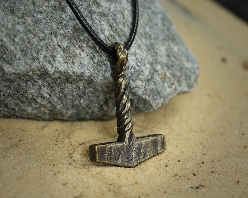 May include: A bronze-colored Mjolnir pendant necklace with a twisted handle, hanging on a black leather cord. The pendant is resting on a light brown sandy surface.