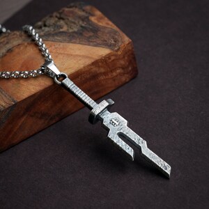 May include: A silver sword pendant necklace with a detailed design. The pendant is attached to a silver chain.