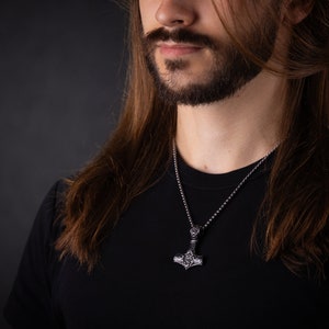 K&ouml;nnte beinhalten: Eine silberne Kettenkette mit einem silbernen Mjolnir-Anh&auml;nger. Der Anh&auml;nger hat komplizierte Details und eine strukturierte Oberfl&auml;che.