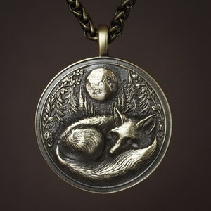 May include: A silver pendant necklace with a detailed design of a sleeping fox under a crescent moon. The pendant is surrounded by a leafy border.