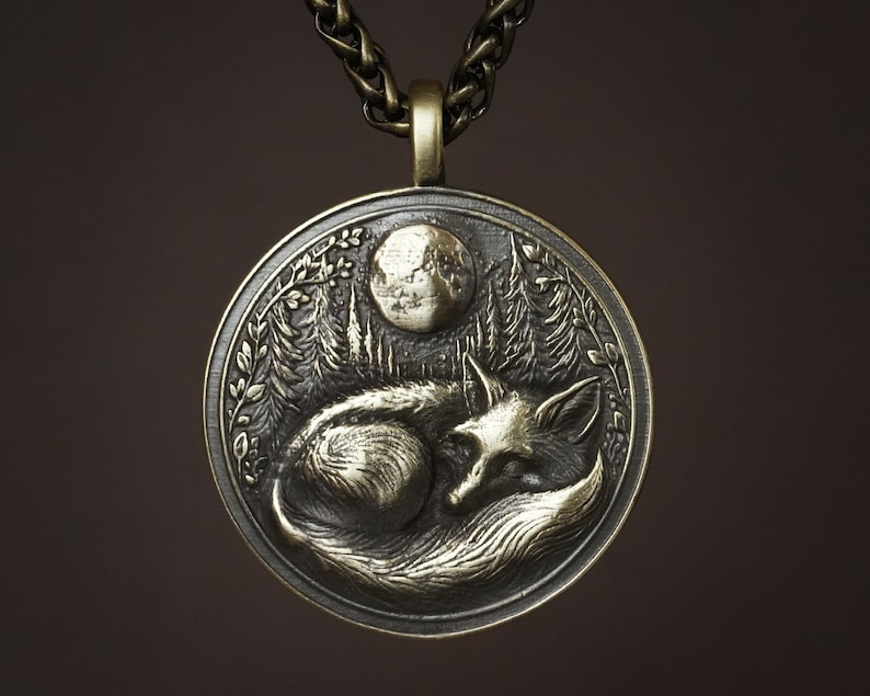 May include: A silver pendant necklace with a detailed design of a sleeping fox under a crescent moon. The pendant is surrounded by a leafy border.