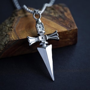 Pu&ograve; includere: Una collana con un ciondolo d'argento che presenta un designo di pugnale dettagliato. Il pugnale ha una punta appuntita e una guardia a forma di croce. La collana &egrave; su una catena d'argento.