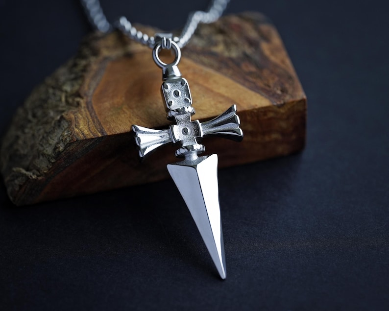 Pu&ograve; includere: Una collana con un ciondolo d'argento che presenta un designo di pugnale dettagliato. Il pugnale ha una punta appuntita e una guardia a forma di croce. La collana &egrave; su una catena d'argento.