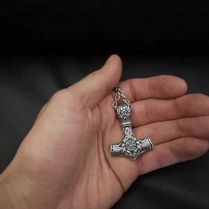 K&ouml;nnte beinhalten: Eine silberne Mjolnir-Halskette mit filigranen Details. Der Anh&auml;nger hat ein kreisf&ouml;rmiges Design mit einem zentralen Stern und ist an einer silbernen Kette befestigt.