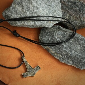 May include: A black leather cord necklace with a gold-toned Mjolnir pendant. The pendant has a detailed design with intricate lines and circles.