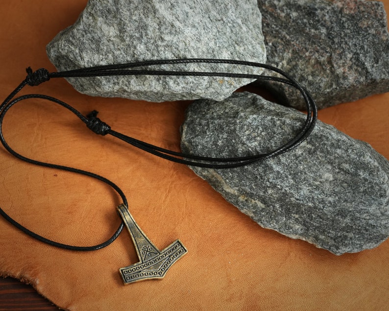 May include: A black leather cord necklace with a gold-toned Mjolnir pendant. The pendant has a detailed design with intricate lines and circles.