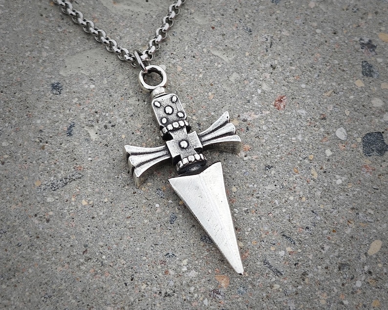Pu&ograve; includere: Una collana con un ciondolo in argento che presenta un design a pugnale dettagliato con un manico a forma di croce.