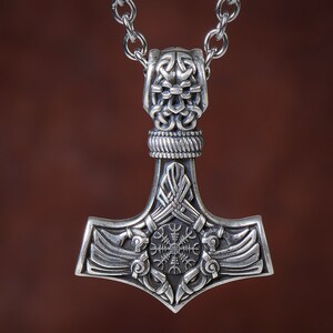 Viking thor hammer pendant showcasing two ravens alongside the Vegvisir, a Viking compass for guidance, and the Aegishjalmur, symbolizing protection. The ravens, intricately detailed, flank the symbols, crafted in silver or bronze