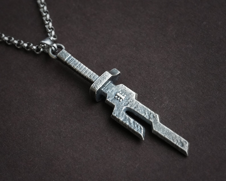May include: A silver sword pendant necklace with a detailed, textured design. The pendant is attached to a silver chain.