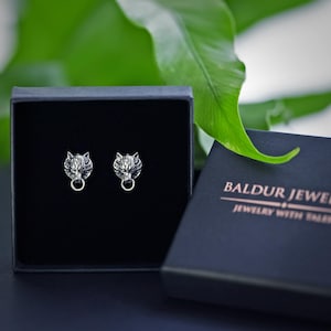 Puede incluir: Un par de pendientes de plata con forma de cabeza de lobo en una caja de joyer&iacute;a negra. La caja tiene el texto "Baldur Jewelry" y "Jewelry with Soul" impreso en ella.