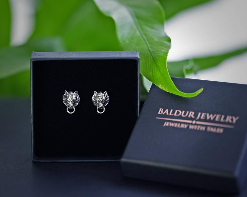 Puede incluir: Un par de pendientes de plata con forma de cabeza de lobo en una caja de joyer&iacute;a negra. La caja tiene el texto "Baldur Jewelry" y "Jewelry with Soul" impreso en ella.