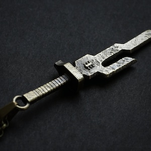 May include: A silver-toned sword pendant necklace with a detailed design. The sword has a textured surface and a pointed tip. The necklace is made of a chain with a lobster clasp.