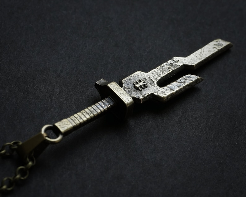 May include: A silver-toned sword pendant necklace with a detailed design. The sword has a textured surface and a pointed tip. The necklace is made of a chain with a lobster clasp.