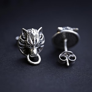 Puede incluir: Un par de pendientes de plata con un dise&ntilde;o de cabeza de lobo. La cabeza de lobo es detallada y tiene un anillo colgando de su boca.
