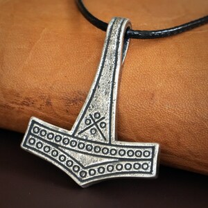 May include: A silver Mjolnir pendant with a black leather cord. The pendant features a detailed design with a cross pattern and a circle border.