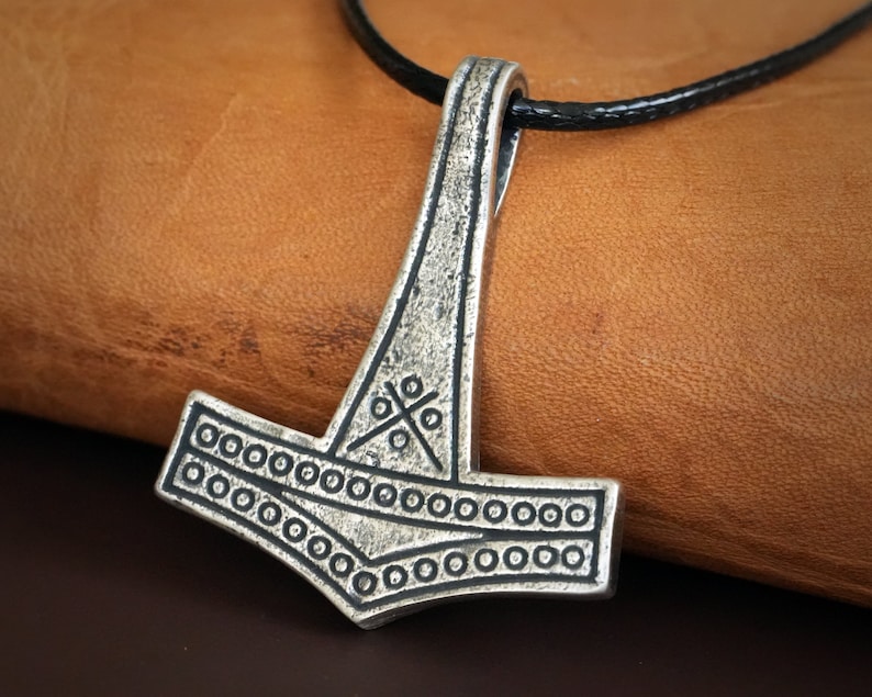 May include: A silver Mjolnir pendant with a black leather cord. The pendant features a detailed design with a cross pattern and a circle border.