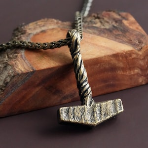 May include: A gold-toned necklace with a twisted chain and a pendant shaped like a hammer. The pendant has a textured surface and a detailed design.