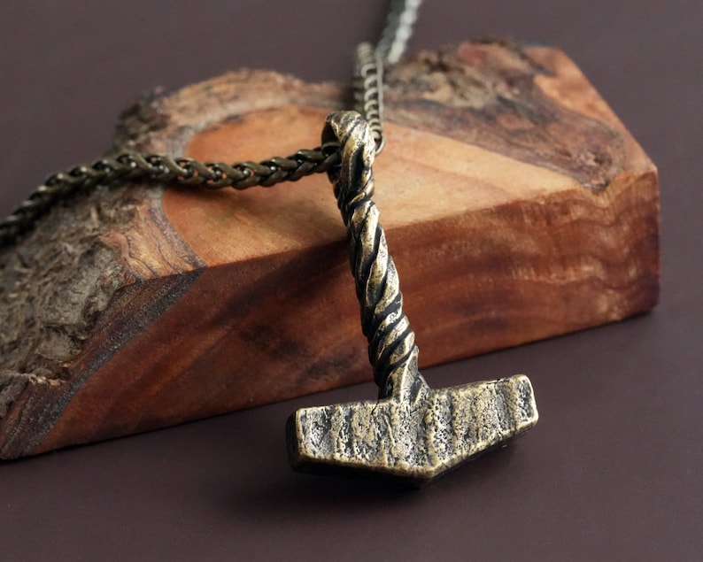 May include: A gold-toned necklace with a twisted chain and a pendant shaped like a hammer. The pendant has a textured surface and a detailed design.