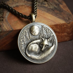 May include: A silver pendant necklace with a detailed design of a fox sleeping under a full moon in a forest setting.