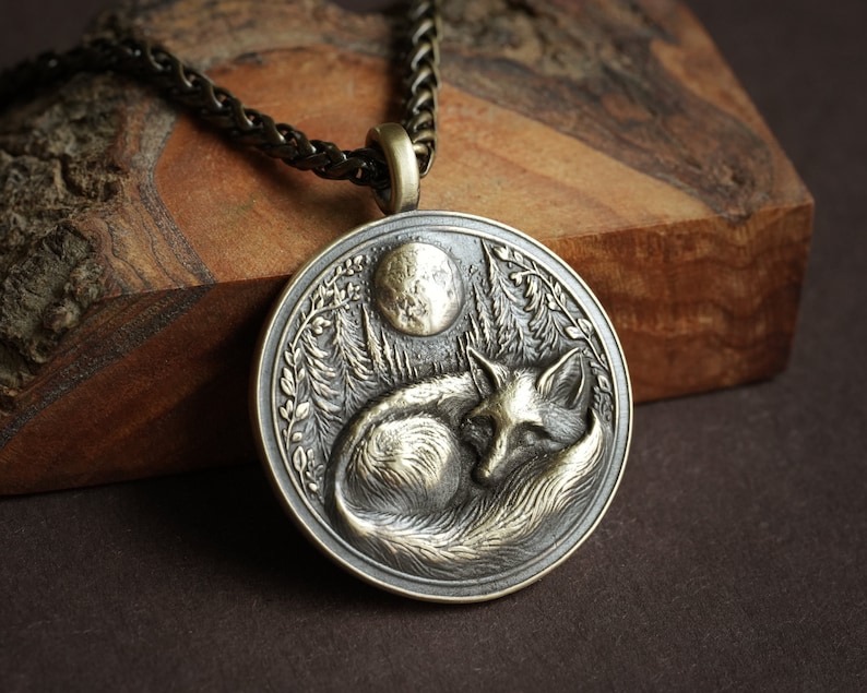 May include: A silver pendant necklace with a detailed design of a fox sleeping under a full moon in a forest setting.