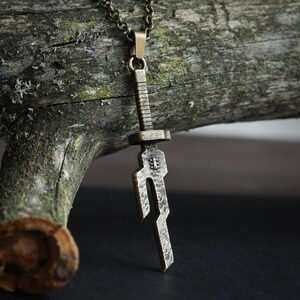 May include: A bronze sword pendant necklace with a detailed design. The pendant is hanging from a chain on a wooden surface.