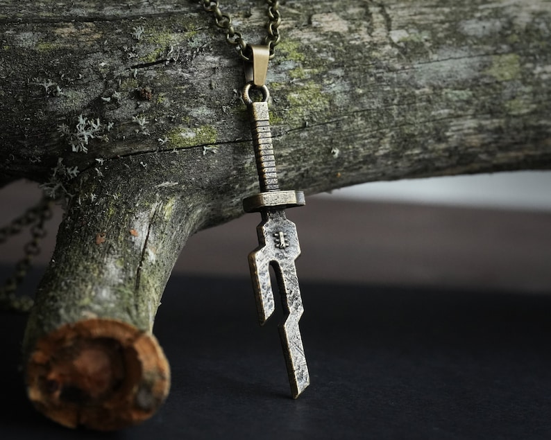 May include: A bronze sword pendant necklace with a detailed design. The pendant is hanging from a chain on a wooden surface.