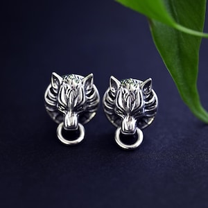 Puede incluir: Un par de pendientes de plata con forma de cabeza de lobo. Los pendientes est&aacute;n detallados con la cara de un lobo y tienen un anillo colgando de la parte inferior.