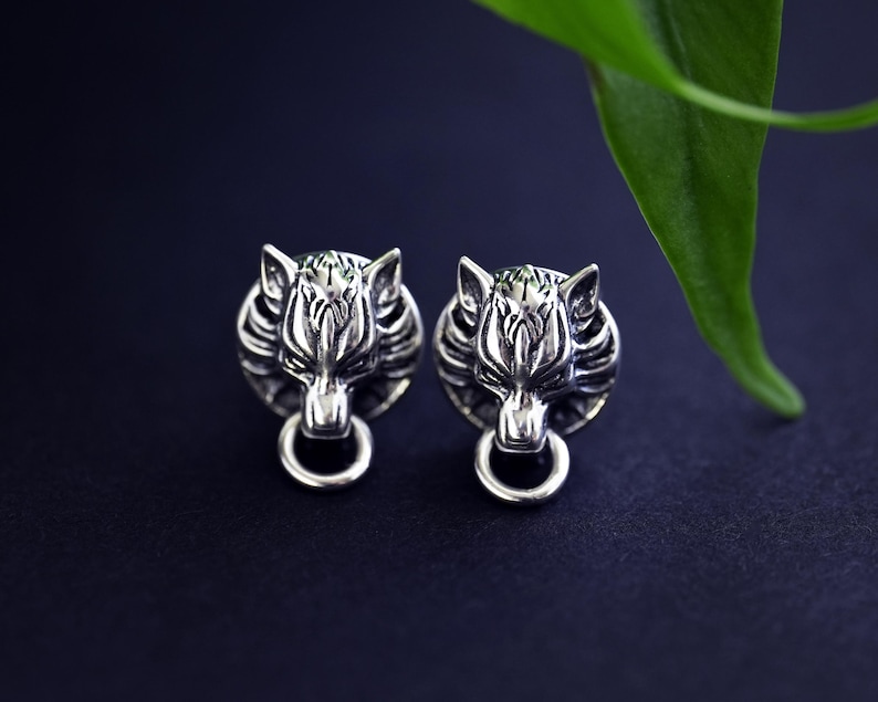 Puede incluir: Un par de pendientes de plata con forma de cabeza de lobo. Los pendientes est&aacute;n detallados con la cara de un lobo y tienen un anillo colgando de la parte inferior.