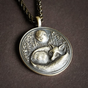 May include: A silver pendant necklace with a detailed design of a fox sleeping under a crescent moon. The pendant is surrounded by a leafy design.