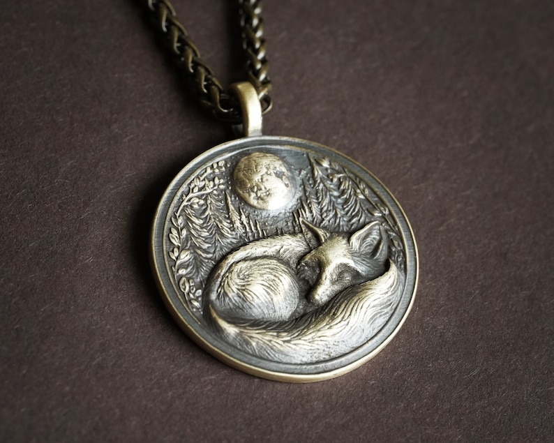 May include: A silver pendant necklace with a detailed design of a fox sleeping under a crescent moon. The pendant is surrounded by a leafy design.