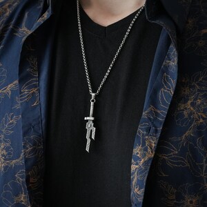 May include: A silver chain necklace with a sword pendant. The pendant is detailed and has a textured finish.