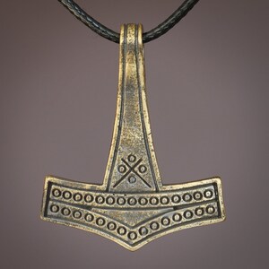 May include: A gold-toned Mjolnir pendant necklace with a black leather cord. The pendant features intricate details, including a circle pattern around the hammer head and a cross design on the top.