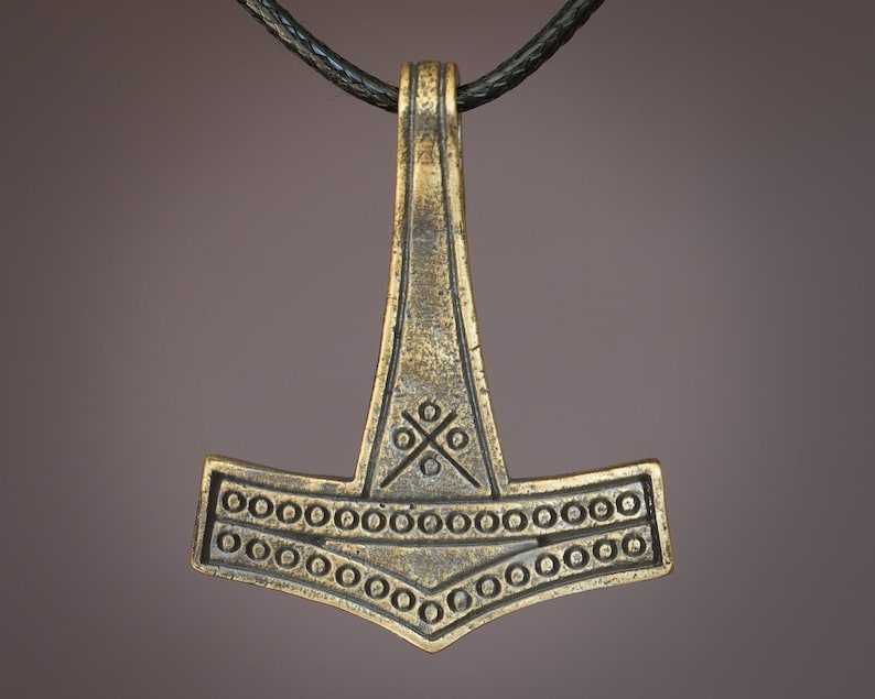 May include: A gold-toned Mjolnir pendant necklace with a black leather cord. The pendant features intricate details, including a circle pattern around the hammer head and a cross design on the top.