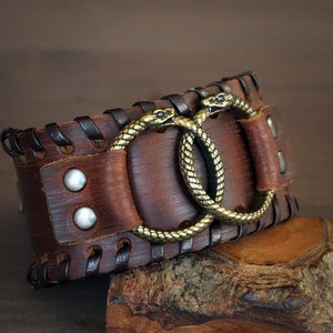 May include: A wide brown leather cuff bracelet with a stitched border and two brass snake head accents. The bracelet is secured with silver-coloured rivets and a leather strap.