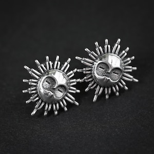May include: A pair of silver-toned stud earrings featuring a sun motif. Each earring displays a central face with closed eyes and a smooth surface, surrounded by a series of radiating, thin, pointed metal rays that extend outward in a circular pattern.