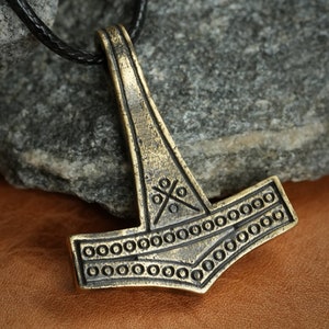 May include: A gold-toned Mjolnir pendant with intricate details, including a cross pattern and a circle border. The pendant is hanging from a black leather cord.