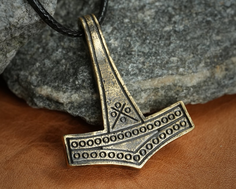 May include: A gold-toned Mjolnir pendant with intricate details, including a cross pattern and a circle border. The pendant is hanging from a black leather cord.