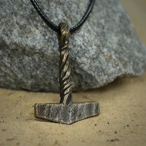 May include: A bronze Mjolnir pendant necklace with a braided rope design on the handle. The pendant is hanging from a black leather cord.