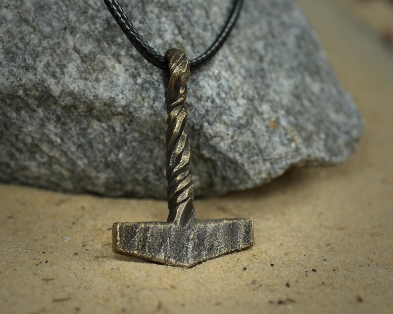 May include: A bronze Mjolnir pendant necklace with a braided rope design on the handle. The pendant is hanging from a black leather cord.