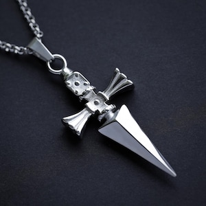 Pu&ograve; includere: Una collana con un ciondolo d'argento a forma di pugnale dettagliato. Il pugnale ha una punta appuntita e un'impugnatura a forma di croce.