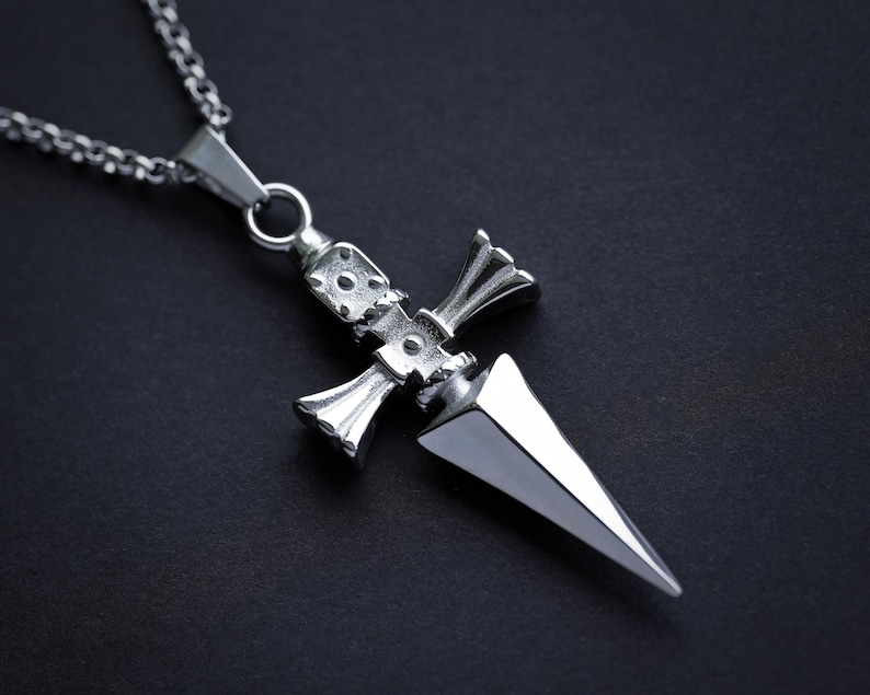 Pu&ograve; includere: Una collana con un ciondolo d'argento a forma di pugnale dettagliato. Il pugnale ha una punta appuntita e un'impugnatura a forma di croce.