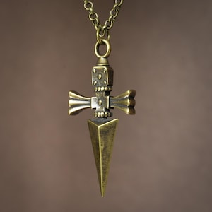 Pu&ograve; includere: Una collana con un ciondolo a forma di pugnale stilizzato con un manico a forma di croce. Il ciondolo &egrave; decorato con intricate incisioni e una punta appuntita.