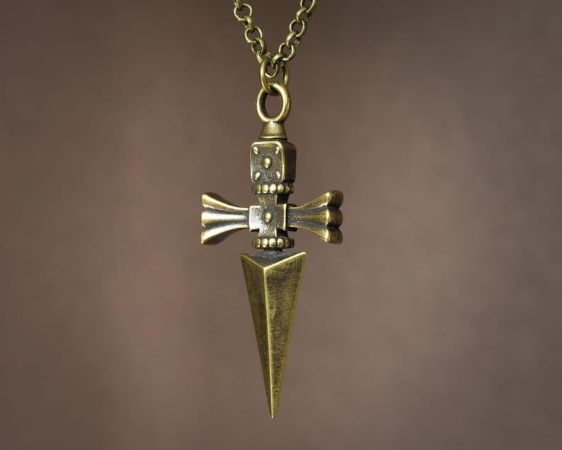 Pu&ograve; includere: Una collana con un ciondolo a forma di pugnale stilizzato con un manico a forma di croce. Il ciondolo &egrave; decorato con intricate incisioni e una punta appuntita.