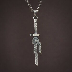 May include: A silver pendant necklace featuring a stylized sword design. The pendant is detailed with intricate lines and a textured finish.