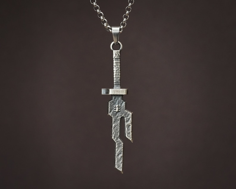 May include: A silver pendant necklace featuring a stylized sword design. The pendant is detailed with intricate lines and a textured finish.