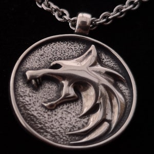 May include: A silver pendant necklace featuring a wolf's head design. The pendant is round and has a textured surface. The wolf's head is facing to the right and has a sharp, pointed snout.
