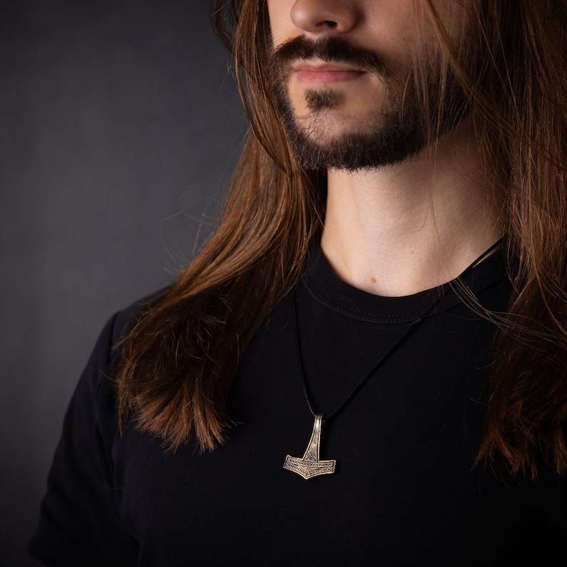 May include: A man with long brown hair and a beard is wearing a black t-shirt with a silver Mjolnir pendant necklace.
