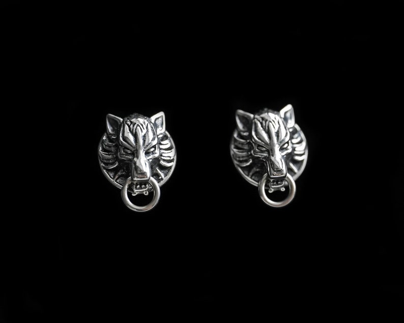 Puede incluir: Un par de pendientes de plata con forma de cabeza de lobo con un detalle de anillo. Los pendientes est&aacute;n sobre un fondo negro.
