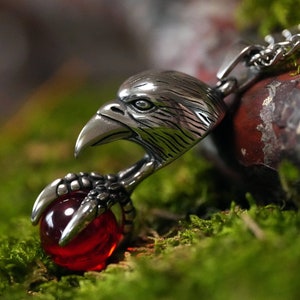 May include: A silver eagle pendant necklace with a red gemstone claw. The eagle's head is facing forward with its beak open. The necklace is made of a silver chain.